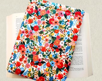 May include: A rectangular fabric sleeve featuring a vibrant floral pattern with red, pink, orange, yellow, and blue flowers interspersed with green leaves on a white background. The sleeve is positioned over an open book with visible text.