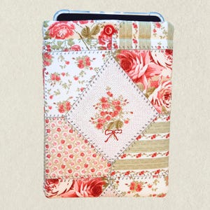 May include: A quilted phone case with a patchwork design. The case features various floral patterns in shades of red, pink, and green on a white and beige background. A central diamond-shaped patch showcases a bouquet of embroidered roses.