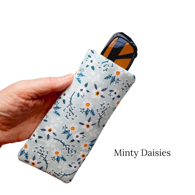 Pu&ograve; includere: Un astuccio per occhiali verde menta con un motivo floreale a margherite, che contiene occhiali da sole. L'astuccio presenta margherite bianche con centri arancioni e foglie e steli blu scuro. Il testo "Minty Daisies" &egrave; visibile.