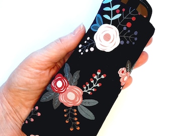Puede incluir: Un estuche de tela negra para gafas con un estampado floral de rosas blancas, rosa claro y rojo oscuro, junto con hojas de color verde oscuro y pequeños detalles en forma de bayas. La funda rectangular tiene esquinas redondeadas y gafas.
