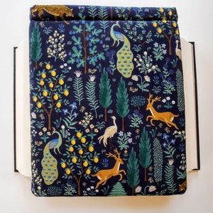 May include: A navy blue book cover with a whimsical forest design. The cover features illustrations of peacocks, deer, lemon trees, and other foliage in shades of yellow, green, and white. The book cover is open, revealing the pages of the book.