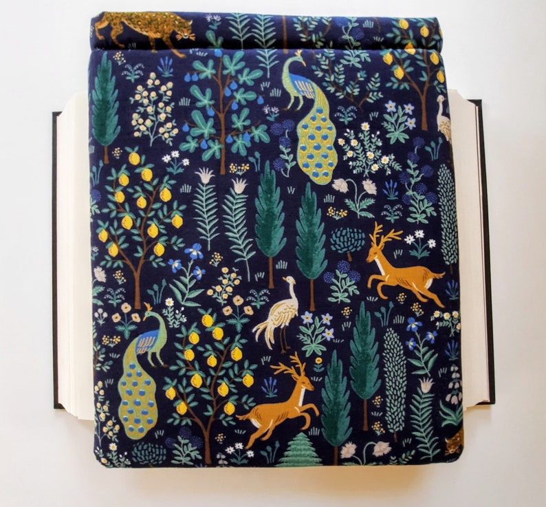 May include: A navy blue book cover with a whimsical forest design. The cover features illustrations of peacocks, deer, lemon trees, and other foliage in shades of yellow, green, and white. The book cover is open, revealing the pages of the book.