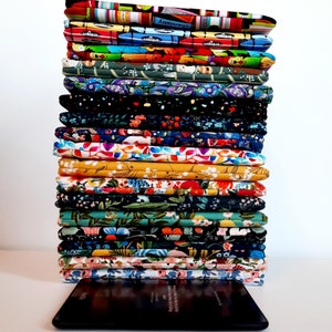 May include: A stack of colorful Kindle sleeves with various patterns, including floral, abstract, and geometric designs. The sleeves are made of fabric and rest on top of a black Kindle device. Text at the bottom reads "Other Kindle sleeves in my shop."