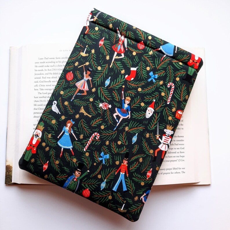 May include: A rectangular fabric book sleeve featuring a black background with a repeating pattern of nutcrackers, ballerinas, candy canes, Christmas ornaments, and green pine branches. The sleeve is folded over at the top edge.