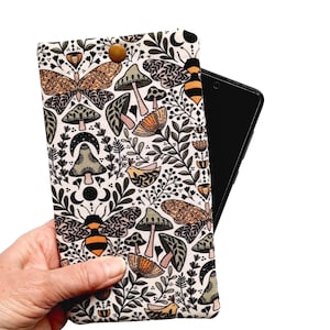 May include: A cream-colored phone case with a whimsical pattern of butterflies, mushrooms, bees, and foliage. The case is held open, revealing a black smartphone. A wooden button secures the case closed.