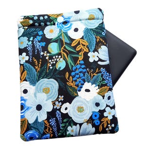 May include: A black floral tablet sleeve with a drawstring closure. The fabric features a pattern of blue and white flowers, green leaves, and gold accents. A black tablet is partially visible inside the sleeve.