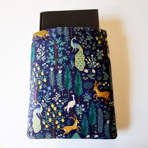 May include: A navy blue book sleeve featuring a whimsical forest design. The fabric is patterned with trees, deer, peacocks, and a leopard. A black book is partially visible inside the sleeve, which is designed to protect books.