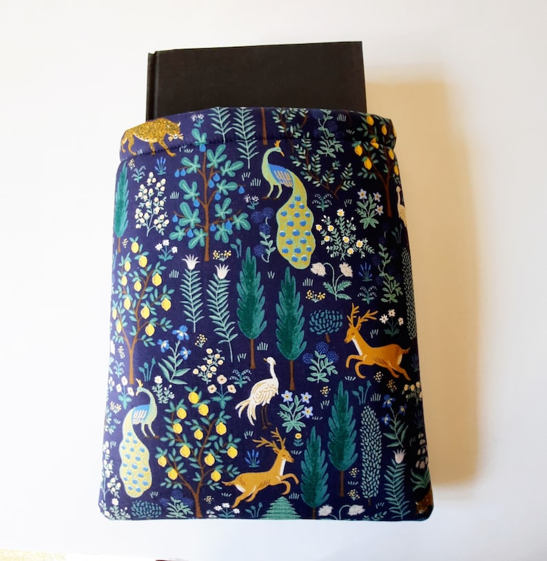 May include: A navy blue book sleeve featuring a whimsical forest design. The fabric is patterned with trees, deer, peacocks, and a leopard. A black book is partially visible inside the sleeve, which is designed to protect books.