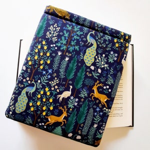 May include: A navy blue book cover with a whimsical forest design. The cover features illustrations of peacocks, deer, and lemon trees. The design is set against a dark blue background with green and yellow accents. The book cover is open, revealing the pages of a book.