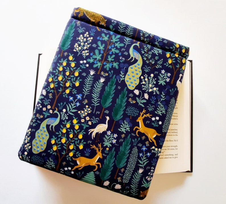May include: A navy blue book cover with a whimsical forest design. The cover features illustrations of peacocks, deer, and lemon trees. The design is set against a dark blue background with green and yellow accents. The book cover is open, revealing the pages of a book.