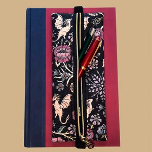 May include: A black pencil case with a gold zipper, featuring a dragon and floral pattern. The case is open, revealing a red and green pen. The case is on top of a red notebook, with a blue book on the left.