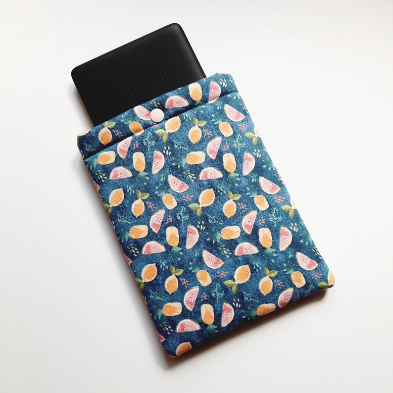 May include: A blue fabric e-reader sleeve with a fruit pattern of lemons and watermelon slices. The sleeve has a button closure and holds a black e-reader. The pattern includes green leaves and is set against a dark blue background.