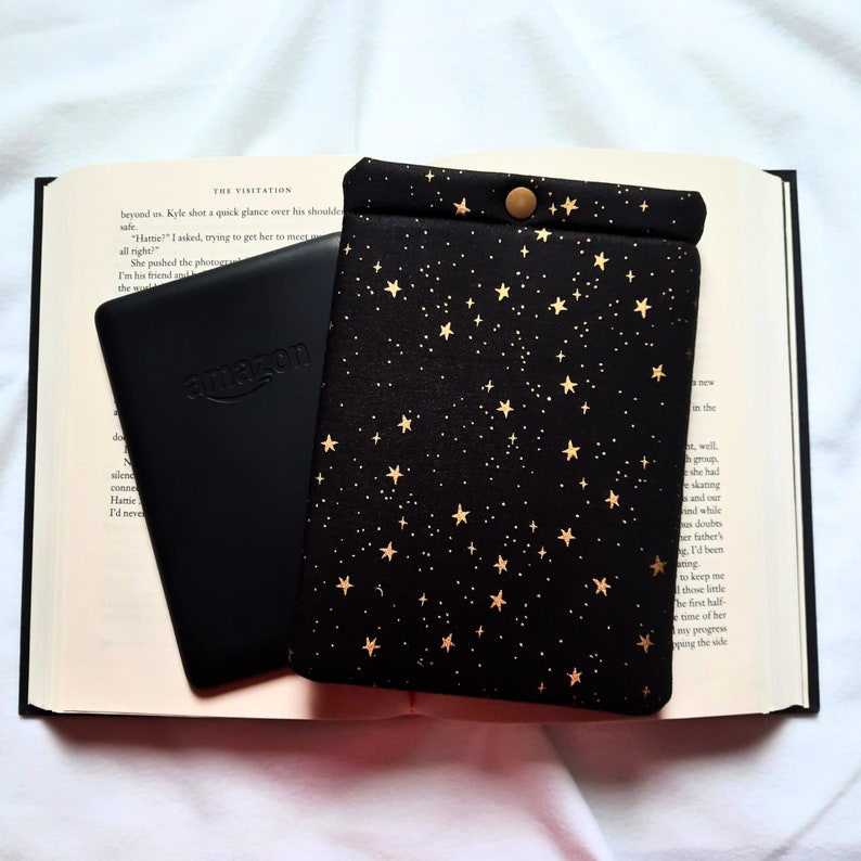 Puede incluir: Un lector electr&oacute;nico negro con la palabra "amazon" en la parte delantera, colocado sobre un libro con una cubierta de patr&oacute;n de estrellas negras y doradas. La cubierta tiene un bot&oacute;n dorado en la parte superior.
