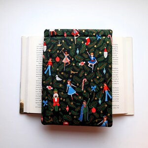 May include: A rectangular fabric book sleeve featuring a dark green background with a repeating pattern of Christmas tree branches, nutcrackers, ballerinas, candy canes, stockings, and ornaments. The sleeve is placed over an open hardcover book.