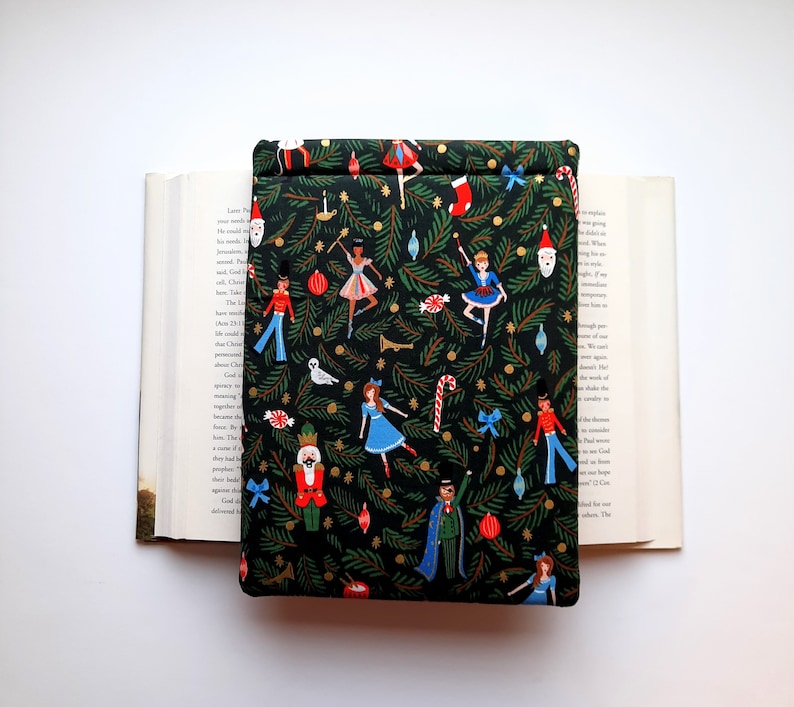 May include: A rectangular fabric book sleeve featuring a dark green background with a repeating pattern of Christmas tree branches, nutcrackers, ballerinas, candy canes, stockings, and ornaments. The sleeve is placed over an open hardcover book.