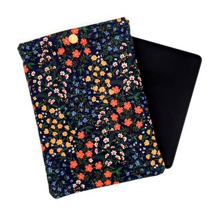 May include: A navy blue floral print e-reader sleeve with a gold button closure. The sleeve features a pattern of orange, yellow, white, and blue flowers. A black e-reader is partially visible.