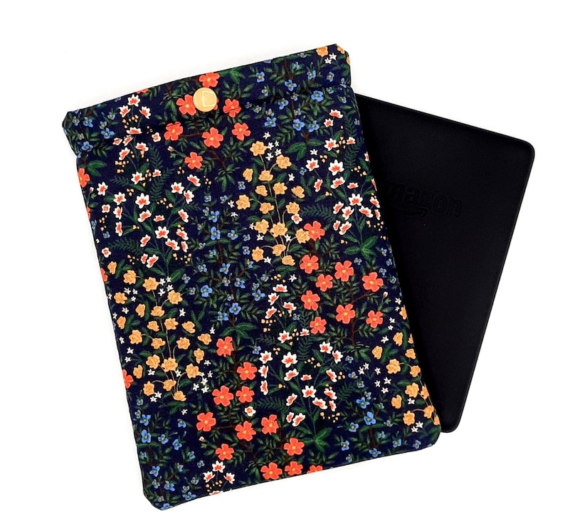 May include: A navy blue floral print e-reader sleeve with a gold button closure. The sleeve features a pattern of orange, yellow, white, and blue flowers. A black e-reader is partially visible.