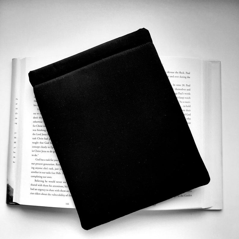 May include: A black book sleeve rests on an open book. The sleeve is rectangular with a soft, matte texture. The book's pages are white, and text is visible. The image is in black and white.