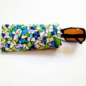 May include: A colorful eyeglasses case with a stained glass pattern in shades of blue, green, and purple. Black sunglasses with orange lenses are partially inserted into the case. The case is rectangular and made of fabric.