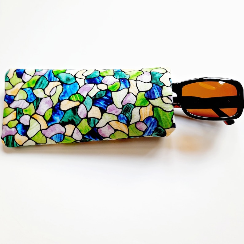 May include: A colorful eyeglasses case with a stained glass pattern in shades of blue, green, and purple. Black sunglasses with orange lenses are partially inserted into the case. The case is rectangular and made of fabric.