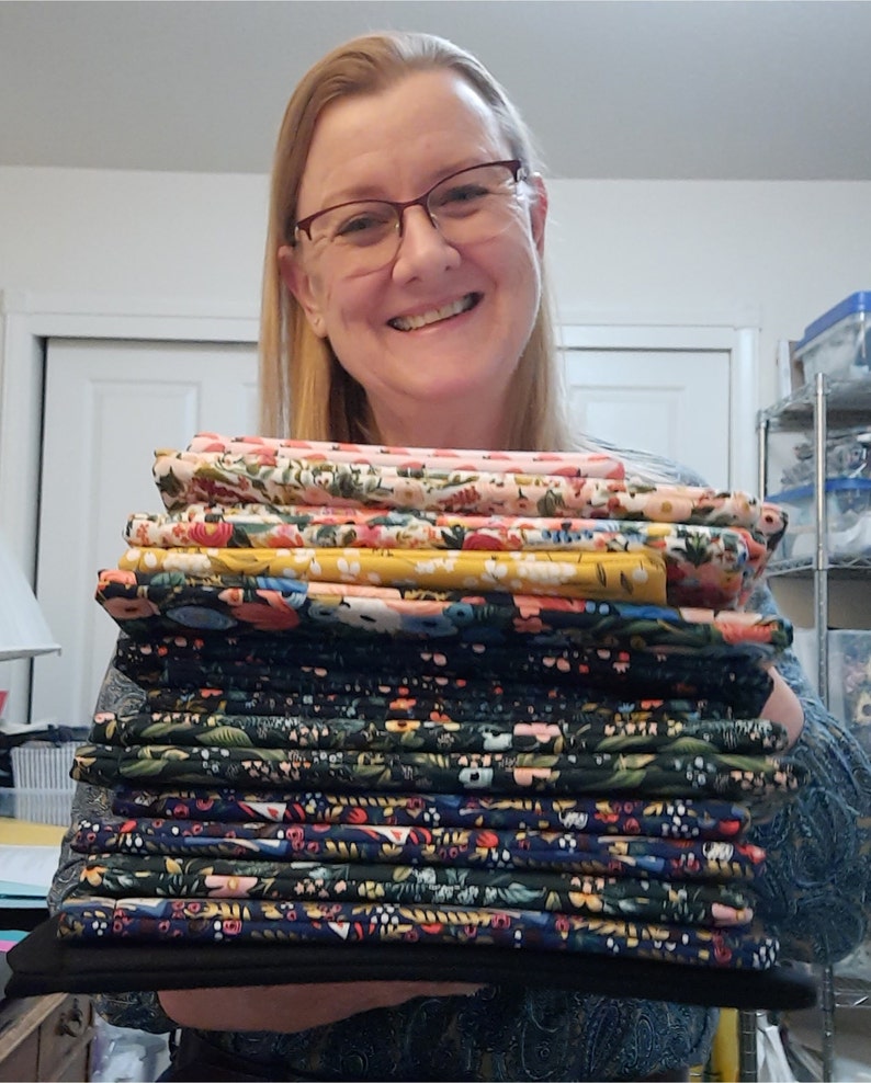 May include: A collection of folded floral fabric in shades of pink, yellow, and navy. The fabrics are held by a person wearing glasses and a patterned top.