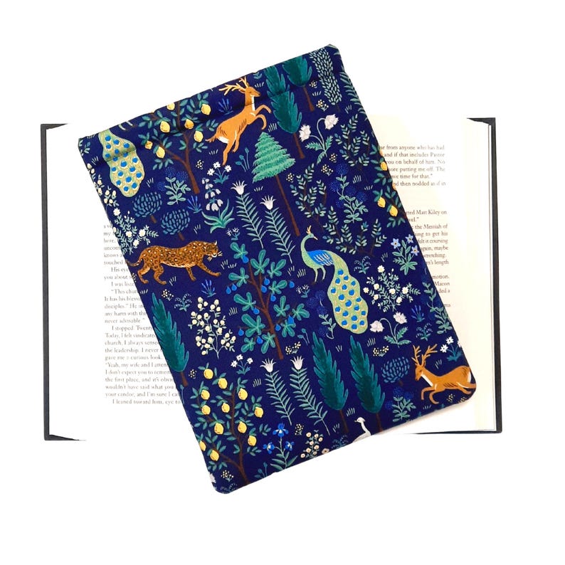 May include: A navy blue book cover featuring a whimsical forest scene. The design includes deer, a leopard, and a peacock amidst trees and foliage. The cover is made of fabric and is placed over an open book.