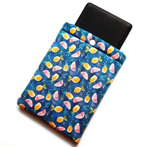 May include: A blue fabric e-reader sleeve with a fruit pattern of lemons and watermelon slices. The sleeve has a button closure and holds a black e-reader with the Amazon logo.
