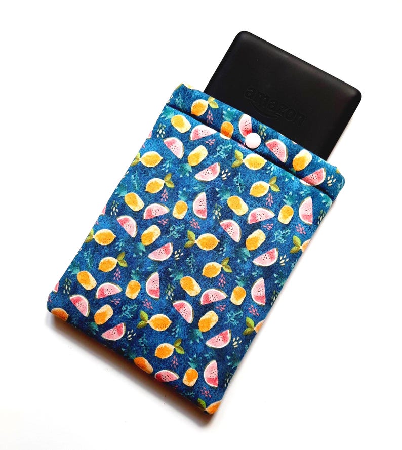 May include: A blue fabric e-reader sleeve with a fruit pattern of lemons and watermelon slices. The sleeve has a button closure and holds a black e-reader with the Amazon logo.