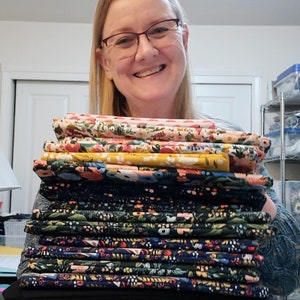 May include: A stack of various floral fabric patterns in pink, yellow, and navy blue. The fabrics are folded and stacked, showcasing a variety of botanical designs. The fabrics are held by a person wearing glasses.