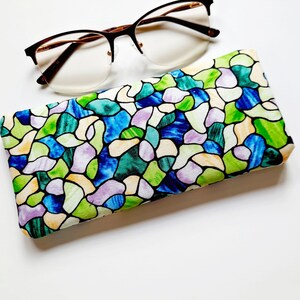 May include: Eyeglasses with a brown frame and a colorful stained-glass pattern glasses case. The case features a design of blue, green, purple, and yellow shapes. The glasses are resting on a white surface.
