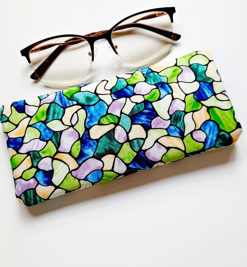 May include: Eyeglasses with a brown frame and a colorful stained-glass pattern glasses case. The case features a design of blue, green, purple, and yellow shapes. The glasses are resting on a white surface.