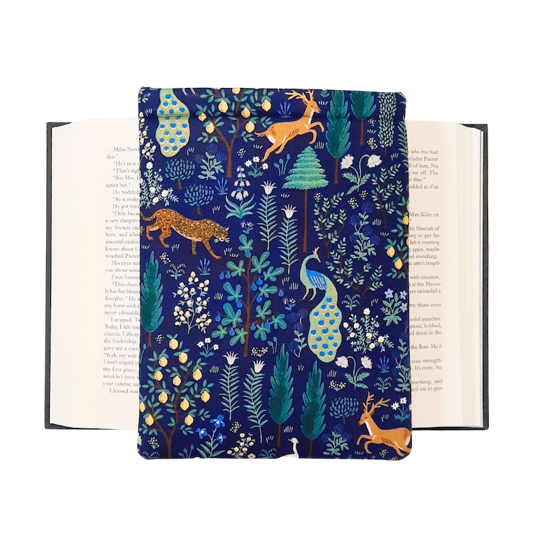 May include: A navy blue book cover with a whimsical forest design. The cover features illustrations of a leopard, deer, and peacock amidst trees and foliage. The cover is open, revealing the pages of a book.