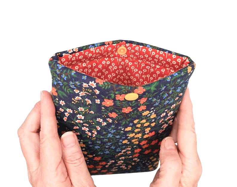 May include: A small pouch with a floral pattern. The exterior is navy blue with a design of colourful flowers. The interior is bright red with a white floral design. It has a yellow button closure.
