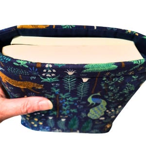 May include: A fabric book sleeve with a dark blue background and a colorful pattern of trees, plants, and animals, including a peacock and a leopard. The sleeve holds two books.