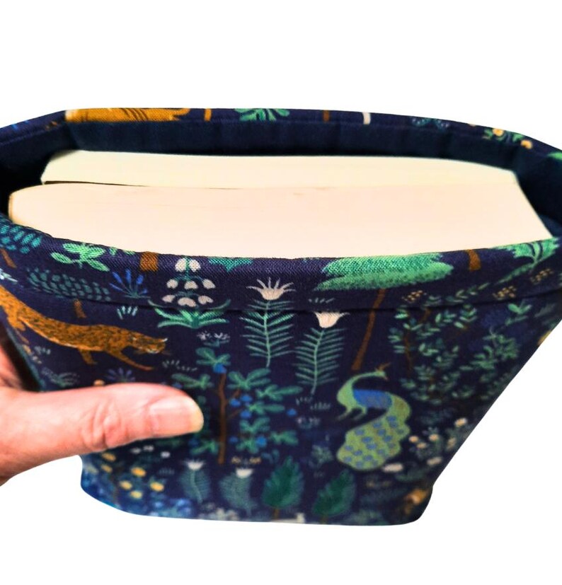 May include: A fabric book sleeve with a dark blue background and a colorful pattern of trees, plants, and animals, including a peacock and a leopard. The sleeve holds two books.