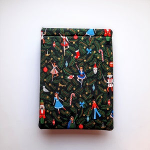 May include: A rectangular fabric pouch featuring a black background with a repeating pattern of green pine branches, red and blue ornaments, candy canes, and figures including nutcrackers, ballerinas, and a bearded man in a red hat.
