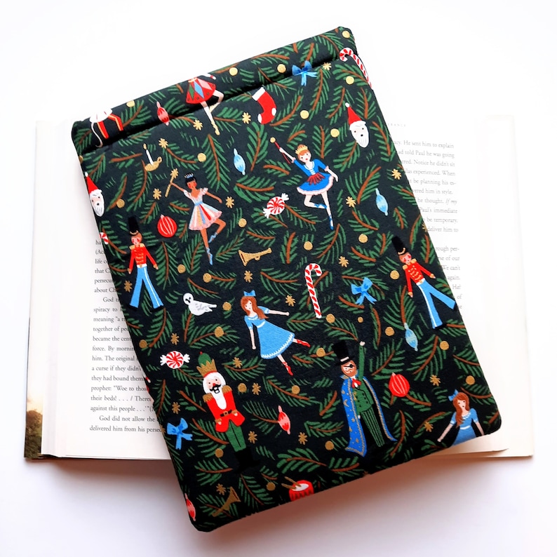 May include: A black fabric sleeve featuring a festive pattern of green pine branches, nutcrackers, ballerinas, candy canes, and holiday ornaments. The rectangular pouch has a folded top edge and is designed to hold books or electronic tablets.