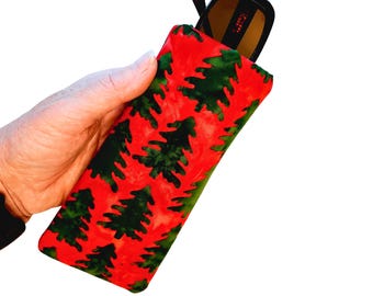 May include: A red eyeglass case with a repeating pattern of green Christmas trees. Black sunglasses are partially visible inside the case. The case is held in a hand against a white background.