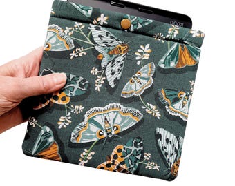 Puede incluir: Una funda de tela verde con un lector electrónico negro en su interior. La funda presenta un patrón repetitivo de polillas y flores coloridas.