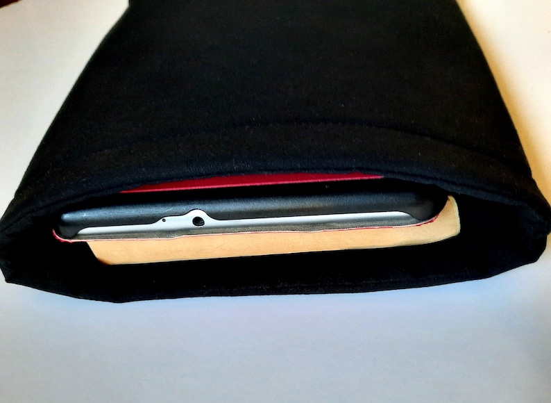 May include: Black fabric sleeve, possibly for a tablet or e-reader, with a device partially visible inside. The device has a black frame, a red border, and a light-colored backing.