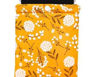 May include: A mustard yellow fabric sleeve for an e-reader featuring a repeating pattern of white floral clusters and thin brown stems. The sleeve has a top flap secured with a single white snap button and contains a black electronic device.
