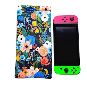 May include: A navy blue floral print glasses case with a button closure next to a Nintendo Switch console. The console has pink controllers on top and green controllers on the bottom.