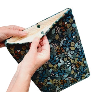 May include: A rectangular, fabric case with a dark background and a botanical print of leaves and flowers in shades of blue, green, and brown. The interior is a light beige color. The case is secured with two green snap closures.