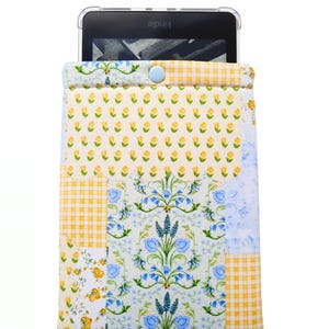 May include: A Kindle e-reader is inside a patchwork fabric sleeve. The sleeve features a floral pattern with yellow, blue, and white colors. The top of the sleeve has a button closure. The word "Kindle" is visible on the e-reader.