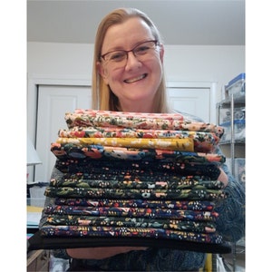 May include: A stack of folded fabric in various floral patterns. The fabrics feature colors like pink, yellow, and navy, with botanical designs. The stack is held by a person wearing glasses and a patterned top.