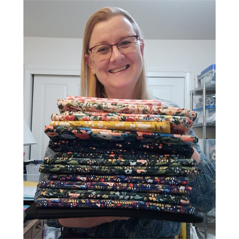 May include: A stack of folded fabric in various floral patterns. The fabrics feature colors like pink, yellow, and navy, with botanical designs. The stack is held by a person wearing glasses and a patterned top.