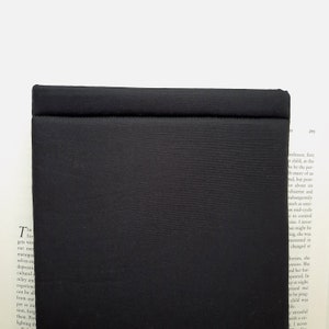 May include: A solid black, rectangular object with a stitched seam across the top. The object has a matte finish and appears to be made of a textured fabric. The item is set against a white background with a book visible in the background.