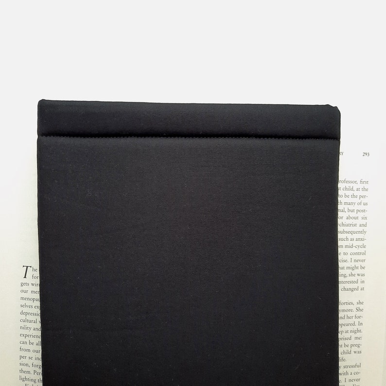 May include: A solid black, rectangular object with a stitched seam across the top. The object has a matte finish and appears to be made of a textured fabric. The item is set against a white background with a book visible in the background.