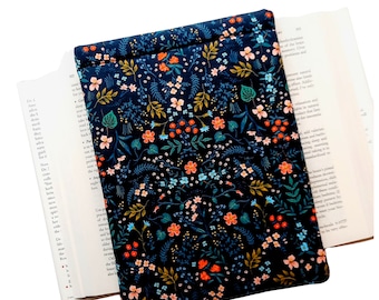 Puede incluir: Una funda rectangular de tela con fondo azul marino oscuro que presenta un denso y colorido estampado botánico de pequeñas flores naranjas, azul claro y rosas, junto con hojas verdes y follaje tipo helecho, colocada sobre un libro abierto.