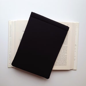May include: A black book cover rests on an open book with text visible on the pages. The book cover is rectangular and covers a portion of the book. The background is white.